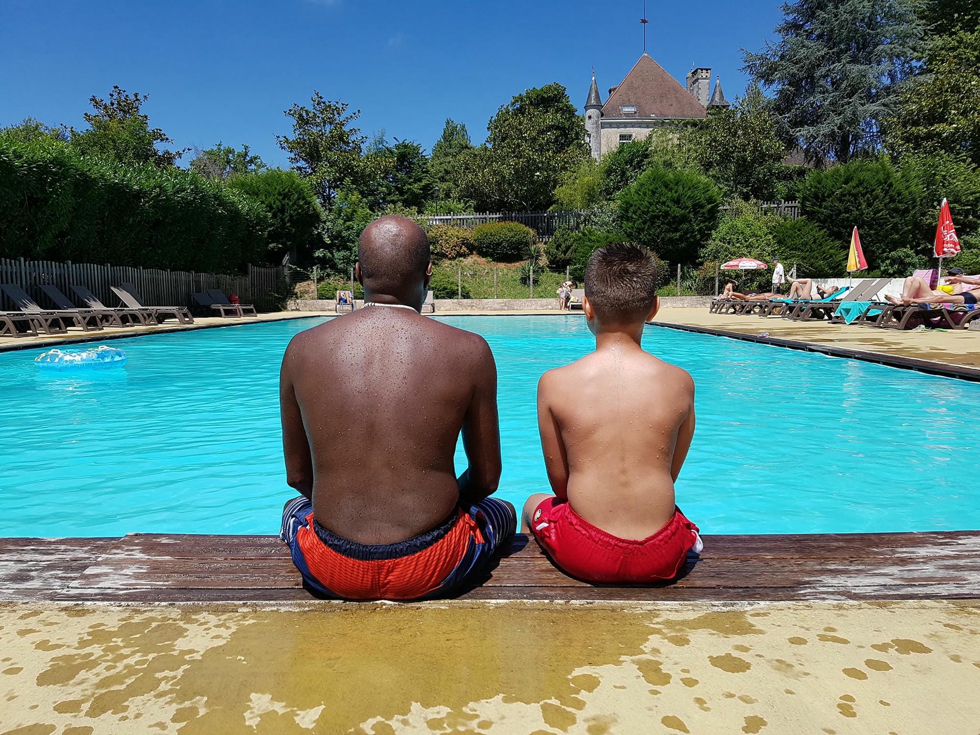 Père et fils assis au bord de la piscine observant l'eau, Camping Le Verdoyer