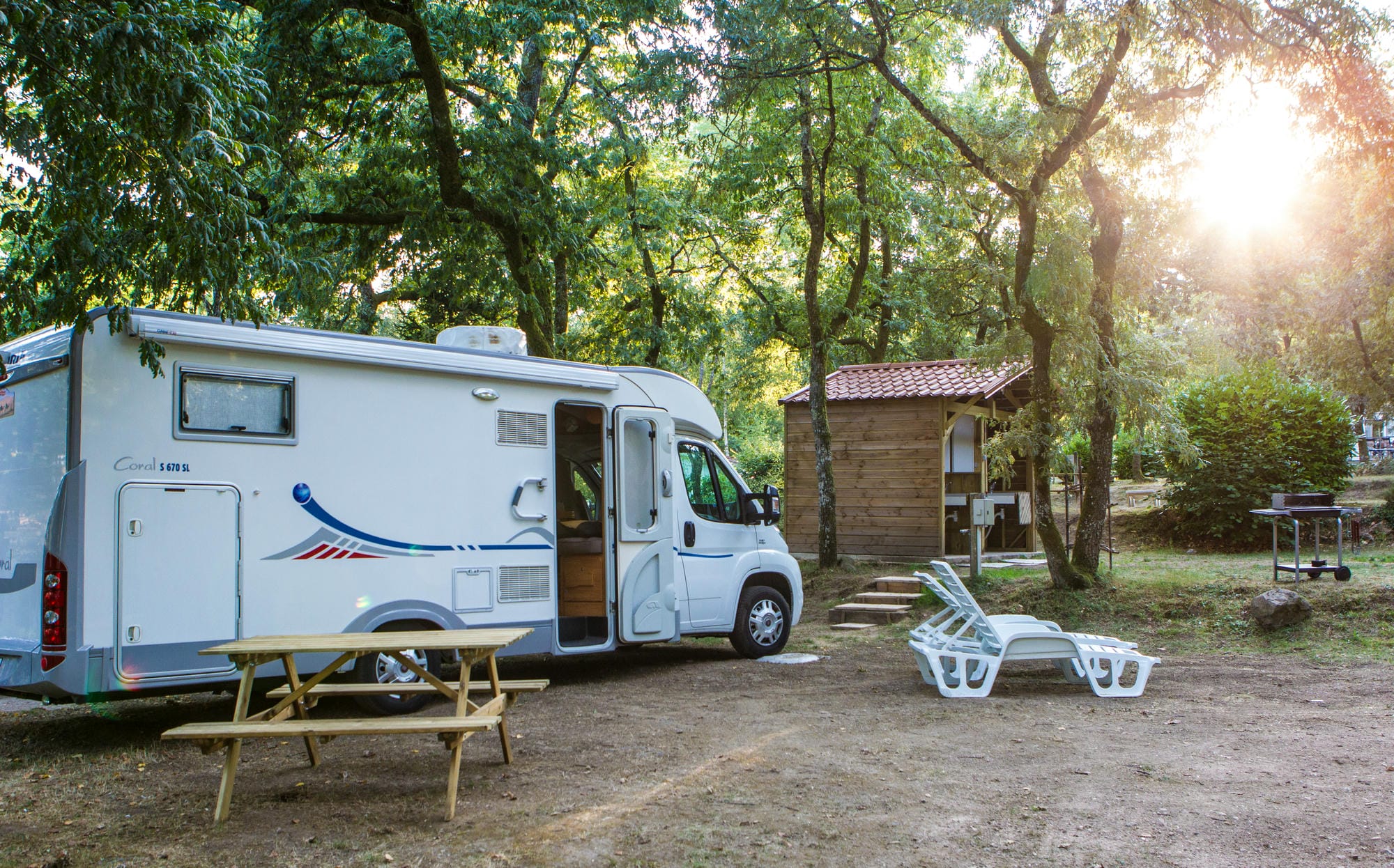 Petit camping-car blanc sur un emplacement boisé avec table de pique-nique au Château le Verdoyer