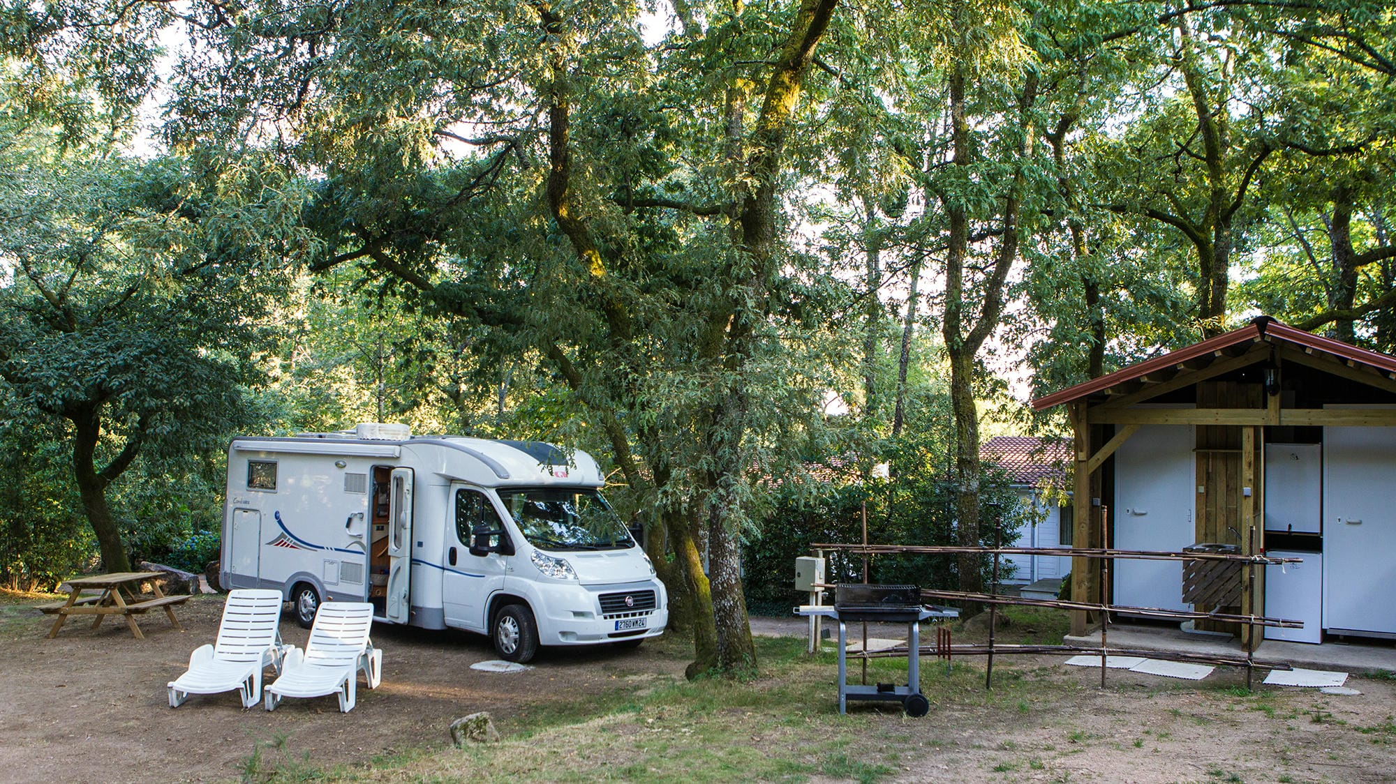 Petit camping-car blanc garé sur un emplacement boisé à côté d'un sanitaire individuel au Château le Verdoyer