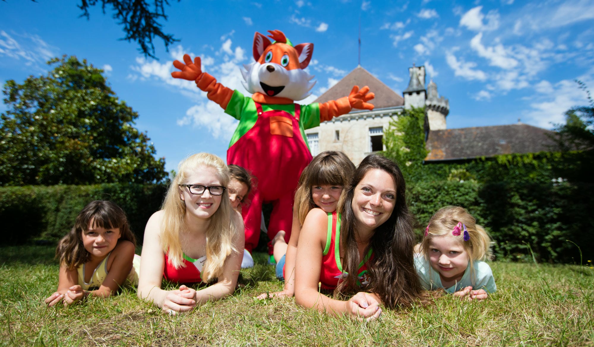 Enfants et Animatrices allongées sur l'herbe devant le château, avec la mascotte Lenny en arrière-plan, club enfants au Verdoyer